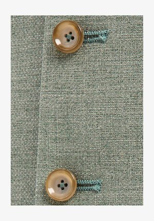 Zwei beige Knöpfe, mit blauem Faden auf strukturiertem grünem Stoff genäht, zeigen Knopflöcher und Stoffdetail.