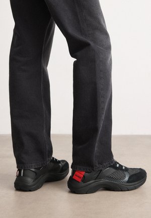Black sneakers with mesh and suede detailing, featuring a red accent tag labeled "HUGO" at the heel, paired with dark gray pants.