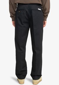 Čierne chinos nohavice so strihom rovného strihu, s dvoma zadnými vreckami a diskrétnym logom na opasku, v kombinácii so svetlo béžovými topánkami.
