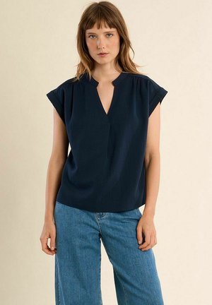 Donna che indossa una canotta blu navy senza maniche con scollo a V e jeans di denim azzurro chiaro su sfondo beige.