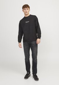 Camiseta negra de manga larga con un pequeño estampado blanco en el pecho, combinada con vaqueros oscuros y zapatillas negras. Diseño minimalista y ajuste casual.