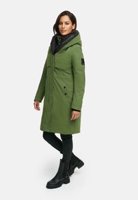 Groene parka met een zwarte capuchon, knopen aan de voorkant en zijzakken. Heeft een gladde textuur en een logo-detail op de capuchon. Zwarte jeans en laarzen.