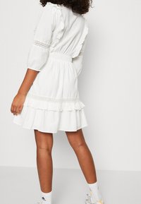 Mujer con un vestido blanco con volantes y detalles de encaje, y mangas abullonadas, combinado con calcetines blancos de Adidas y zapatillas blancas, de pie contra una pared lisa.