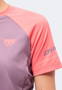 Gros plan d’une personne portant un maillot de sport rose et violet avec un logo de panthère sur la poitrine et le texte "DYNA" sur la manche.