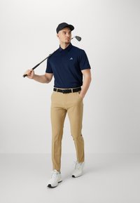 Marinblå pikétröja med korta ärmar, tanfärgade byxor, svart bälte och vita träningsskor. Håller ett golfklubba, bär en svart keps.