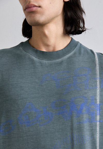 T-shirt en coton vert avec une finition texturée, arborant des motifs de texte graphique bleu. Encolure ronde et détail de couture contrasté sur le côté.