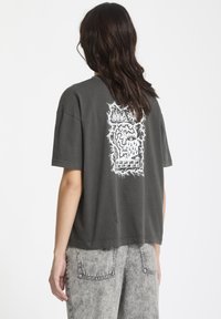 Camiseta de gran tamaño en gris oscuro con un diseño gráfico blanco en la parte posterior. El tejido parece suave, con un corte relajado y mangas cortas.