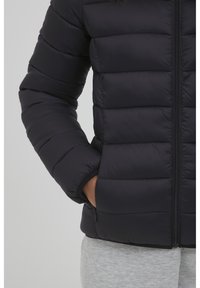 Veste noire rembourrée avec fermeture éclair à l'avant, dotée d'un design matelassé et de poches latérales. Les manches sont ajustées avec une texture lisse.