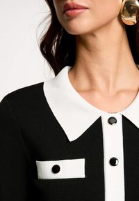 Prenda con cuello en blanco y negro que presenta una textura acanalada, botones negros y un pequeño bolsillo blanco con un detalle de botón negro.