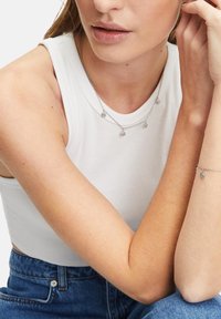Weißes geripptes Tanktop, silberne mehrlagige Halskette mit kleinen Anhängern und ein zartes Armband, getragen mit hoch taillierten blauen Jeans.