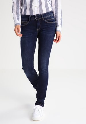 Personne portant un jean slim bleu foncé, un t-shirt à manches longues blanc et gris rayé, et des baskets blanches à lacets.