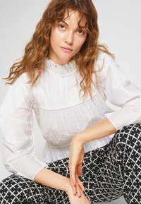 Vit plisserad blus med hög volangkrage och genomskinliga ärmar, i kombination med svarta och vita mönstrade byxor med geometrisk design.