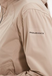 Veste imperméable beige avec une texture douce, dotée d'un design ajusté et d'un logo "ENDURANCE" brodé sur la manche gauche.