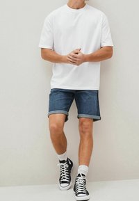 Biała koszulka z krótkim rękawem w połączeniu z ciemnoniebieskimi szortami dżinsowymi z podwiniętymi nogawkami oraz czarnymi sneakersami za kostkę z białą podeszwą i skarpetkami do kostek.