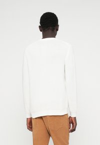 Λευκό πουλόβερ με ρίγες, στρογγυλή λαιμόκοψη και μακριά μανίκια, συνδυασμένο με καμηλό παντελόνι από κόρντουρα, προβαλλόμενο από πίσω.