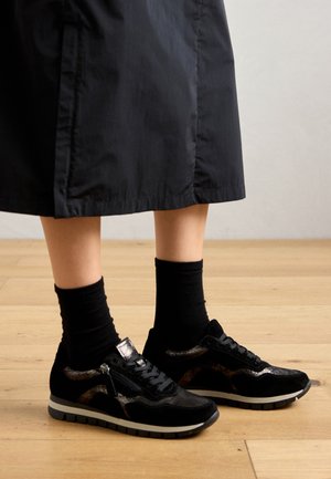 Sneaker in suede nera con accenti metallici, chiusura con lacci e suola grigia spessa, abbinate a calzini neri a crew e una gonna scura.