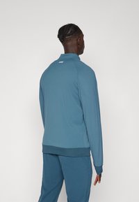 Jachetă sport teal cu guler înalt, mâneci lungi și tiv cu nervuri. Prezintă o textură netedă cu detalii de cusătură tonale subtile.
