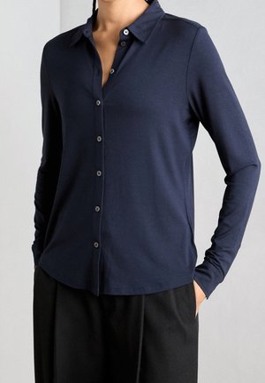 Personne portant une chemise bleu marine à manches longues boutonnée, associée à un pantalon noir, debout les mains dans les poches sur un fond uni.