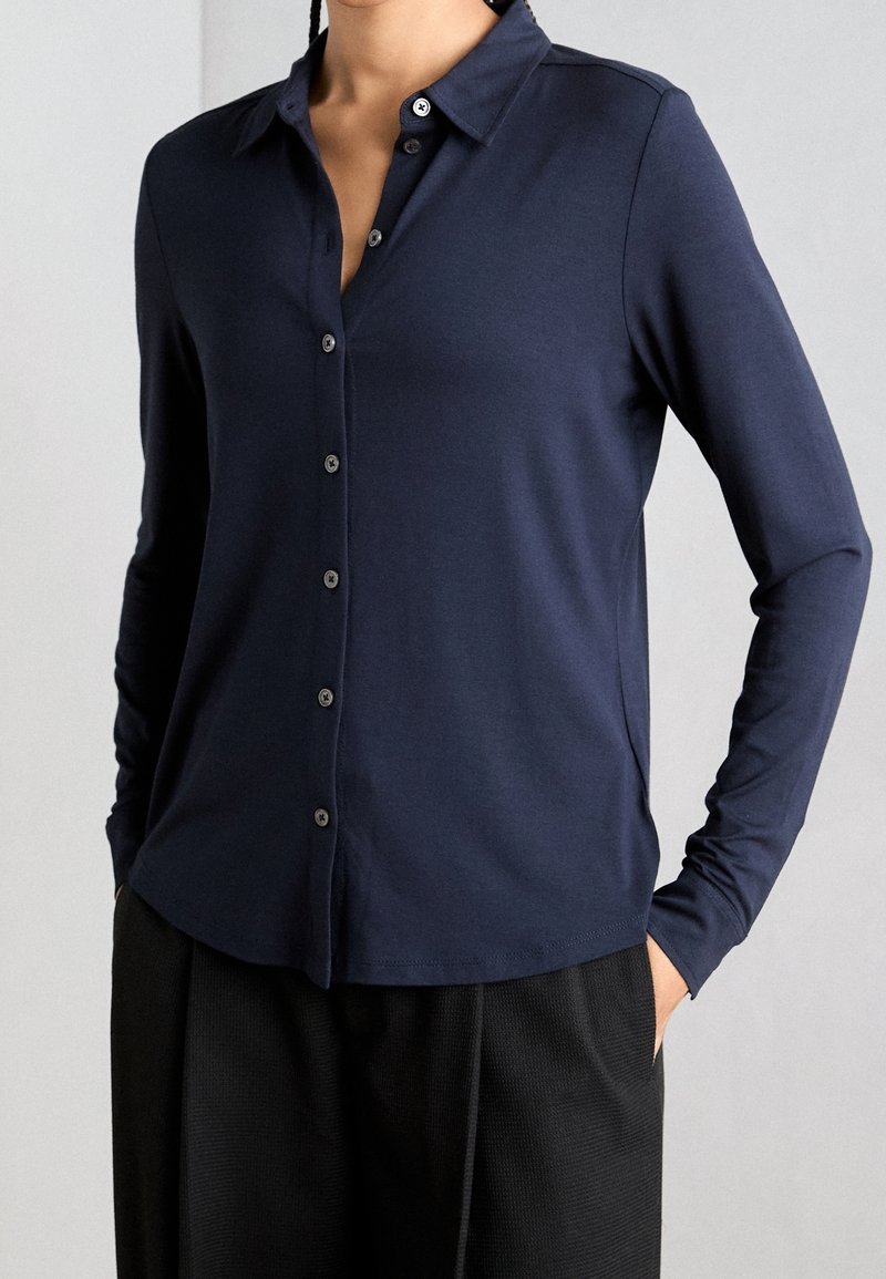 Personne portant une chemise bleu marine à manches longues boutonnée, associée à un pantalon noir, debout les mains dans les poches sur un fond uni.