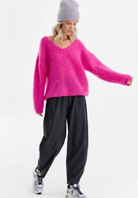 Pulover roz aprins, moale și supradimensionat, cu decolteu în V, asortat cu pantaloni negri cu dungi subțiri și pantofi sport albi. Textură tricotată, croială lejeră.