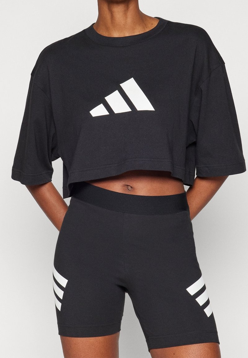 Černé zkrácené tričko s bílým logem se třemi pruhy na přední straně, doplněné černými šortkami s bílými pruhy po stranách.