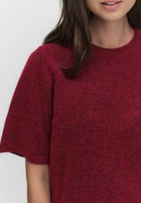 Bordowy sweter z krótkim rękawem o miękkim, teksturowanym splocie. Okrągły dekolt i luźny krój, podkreślający minimalne detale szwów.