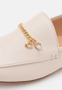 Chaussure en cuir beige clair à texture lisse, ornée d'une chaîne dorée et d'un pendentif. Bout arrondi et surpiqûres le long du bord.