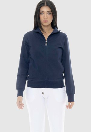 Donna che indossa una giacca blu navy con zip e tasche laterali e pantaloni bianchi, in piedi davanti a uno sfondo grigio chiaro uniforme.