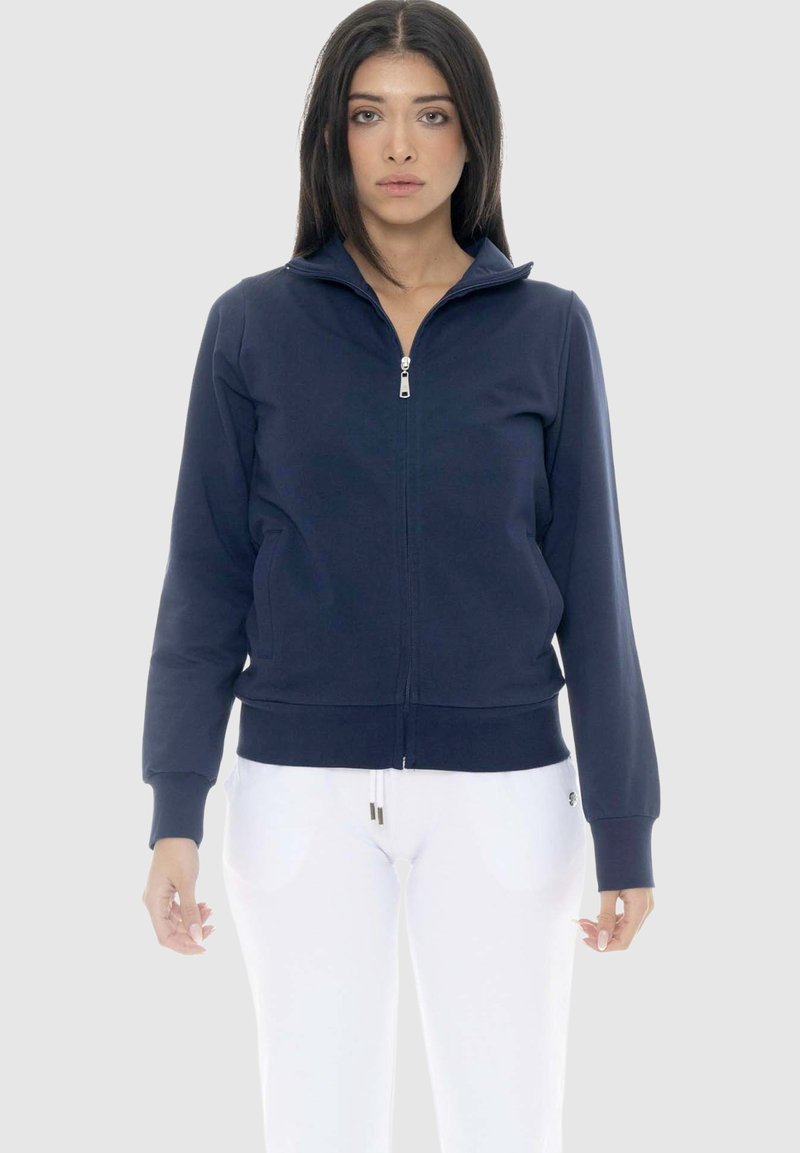 Femme portant une veste zippée bleu marine avec des poches latérales et un pantalon blanc, debout devant un fond gris clair uni.