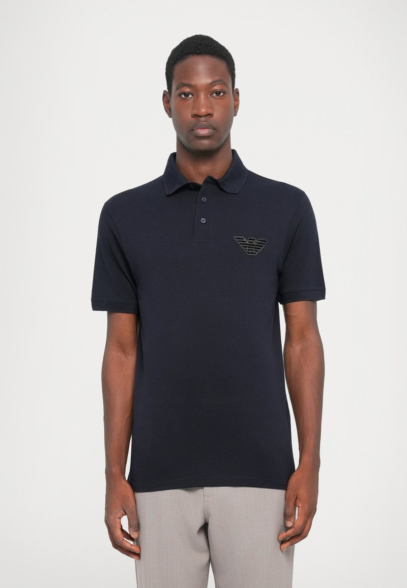 Tumši zilā kokvilnas polo krekls ar īsām piedurknēm, ar trīs pogu aizdari un izšūtu emblēmu kreisajā krūtī.