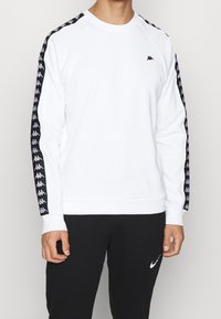 Witte sweatshirt met zwart Kappa-logo op de borst en zwarte gestreepte zijstrips. Beschikt over een geribde halslijn en manchetten. Gepaarde met zwarte broek.