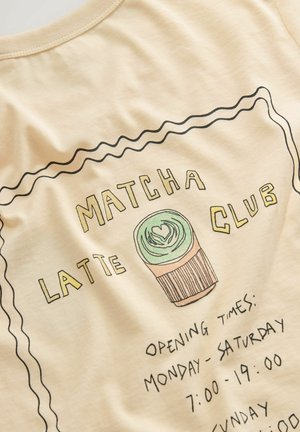 Kremowa koszulka z graficznym wzorem zawierającym napisy "MATCHA LATTE CLUB", ilustrację babeczki oraz godziny otwarcia napisane czarnym atramentem.