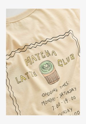 Kremo spalvos marškinėliai su grafiniu dizainu, kuriame yra užrašas "MATCHA LATTE CLUB", cupcakes iliustracija ir darbo valandos juodu rašalu.