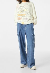 Felpa oversize crema con testo e design grafico, abbinata a pantaloni cargo in denim blu a gamba larga con tasche laterali. Calzature casual visibili.