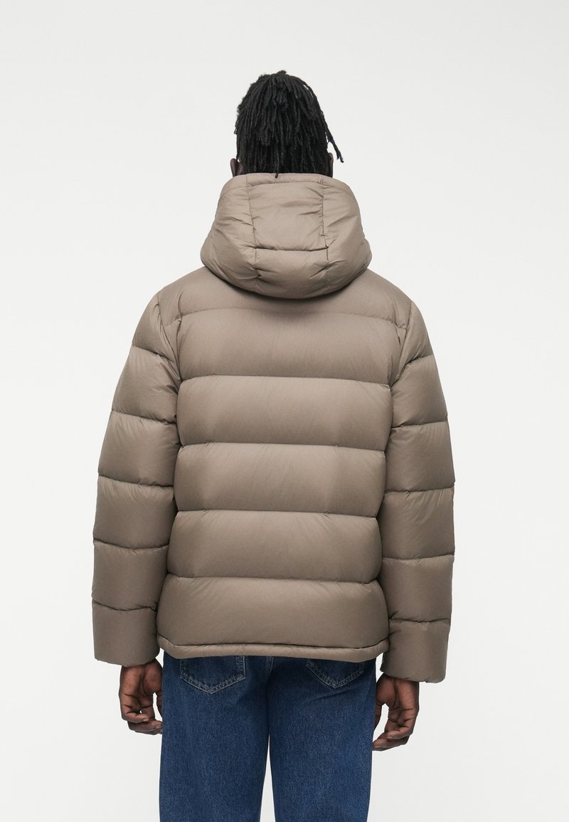 Person mit dunklen geflochtenen Haaren, die eine taupefarbene, wattierte Kapuzenjacke und blaue Jeans trägt, steht mit dem Rücken zur Kamera vor einem einfarbigen Hintergrund.