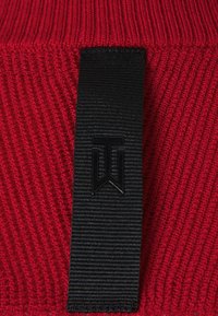 Tissu tricoté rouge avec une étiquette tissée noire verticale comportant un logo en relief embossé avec des lettres stylisées "T" et "W".