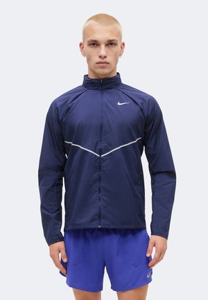 Homme aux cheveux blonds courts portant une veste zippée Nike bleu marine et un short Nike bleu, debout devant un fond clair uni.