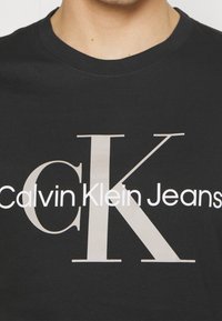 T-shirt in cotone nero con grande logo "CK" bianco e beige chiaro, accompagnato dalla scritta "Calvin Klein Jeans" sottostante in carattere grassetto.