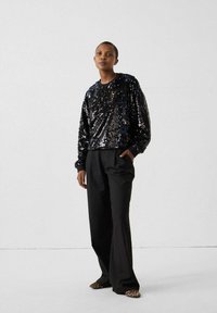 Vrouw staat tegen een witte muur, draagt een zwarte top met lange mouwen en pailletten, losse zwarte broek en puntige schoenen met luipaardprint.