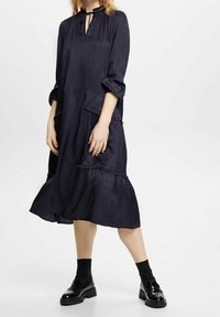 Mörkblå satin-klänning med nyckelhålskrage, långa ärmar, fransad nederdel och två sidfickor; mjuk textur och flödande silhuett.