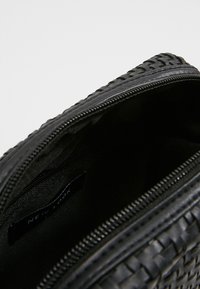 Sac à main tissé noir avec une fermeture éclair. L'extérieur présente une surface texturée, tandis que l'intérieur est en tissu noir uni. Étiquette : "NEW LOOK."