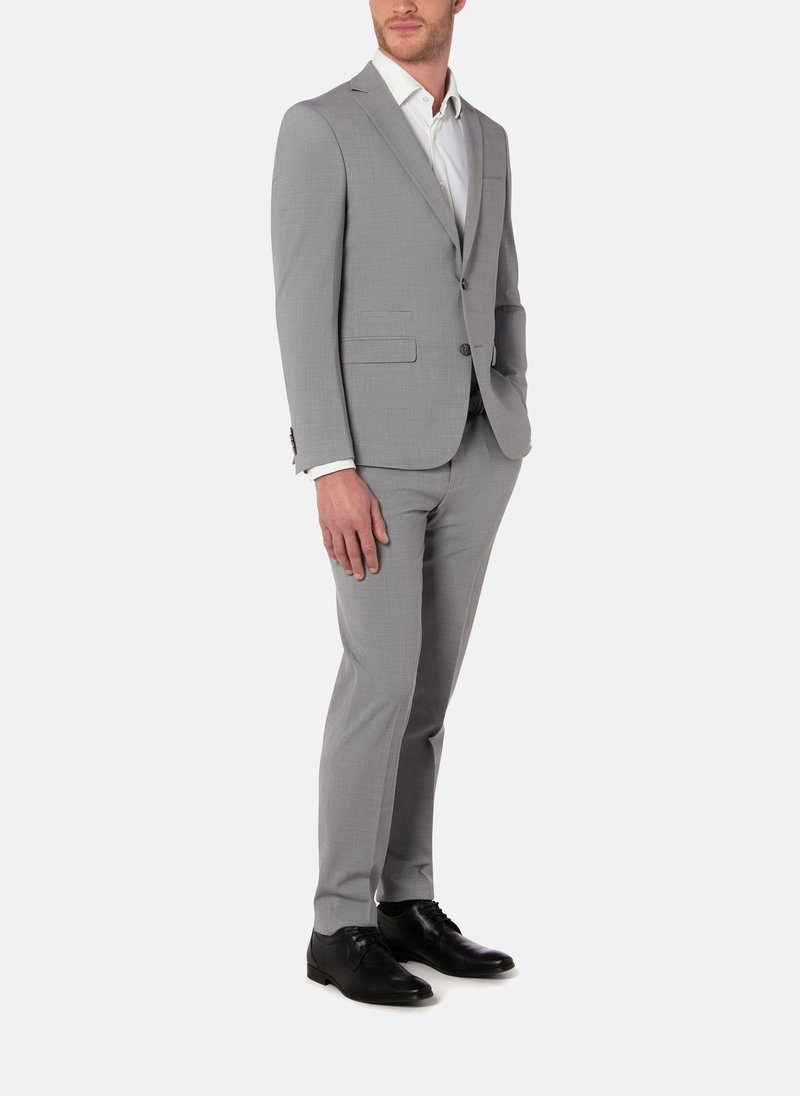 Homme portant un costume slim gris clair avec une chemise blanche et des chaussures noires, debout devant un fond blanc.
