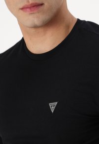 Svart bomull T-skjorte med rund hals og et lite trekantet logo-patch på brystet med "GUESS" i hvit tekst.