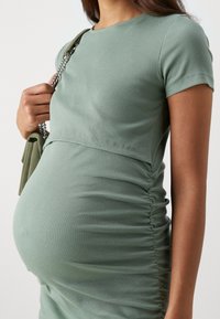 Umstandskleid aus sanftem grünen Rippstoff, mit kurzen Ärmeln und gerafften Details an der Seite für eine taillierte Silhouette.