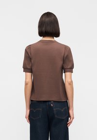 Femme aux cheveux courts et foncés, portant un haut marron à manches courtes et un jean bleu foncé, debout de dos sur un fond uni.