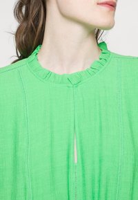 Blouse en tissu vert texturé avec un col à volants et des détails de coutures verticales. Comprend une ouverture en forme de goutte d'eau à l'avant.