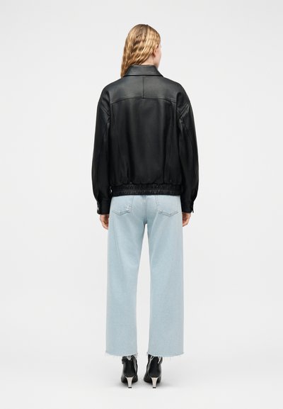 Γυναίκα με μακριά ξανθά μαλλιά που φοράει μαύρο δερμάτινο μπουφάν, ανοιχτό μπλε τζιν με φαρδιές παντελόνες και μαύρα μποτάκια με τακούνι, γυρισμένη με την πλάτη.