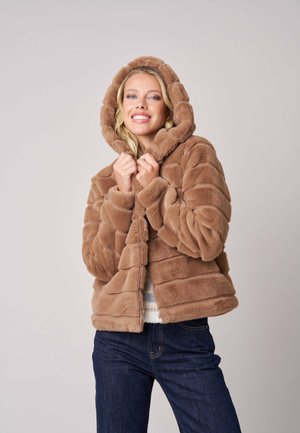 Femme souriante portant une veste à capuche marron duveteuse et un jean bleu foncé, tenant le col de la veste avec les deux mains.