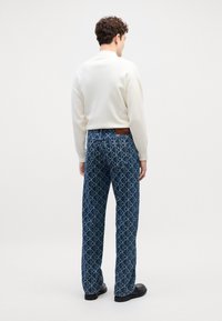 Jeans azules con un diseño geométrico, combinados con un suéter ligero color crema. Los jeans tienen una etiqueta de cuero marrón en la parte trasera.