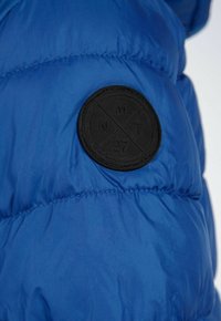 Sektion med blå puffjacka som har ett texturerat svart gummiloggopatch med cirkeldesign, synliga sömmar och en slät, reflekterande tygyta.